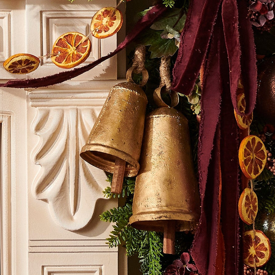A festive Christmas mantel decorated with a green garland featuring eucalyptus, berries, dried orange slices, gold bells, and metallic ornaments, with a framed painting of Santa Claus above a white fireplace and lit candles around it.