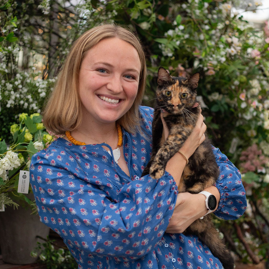 A smiling woman with blonde hair holds a tortoiseshell cat in a lush greenhouse or garden center. She is wearing a blue long-sleeve blouse with a small pink and white floral pattern, paired with light blue jeans.