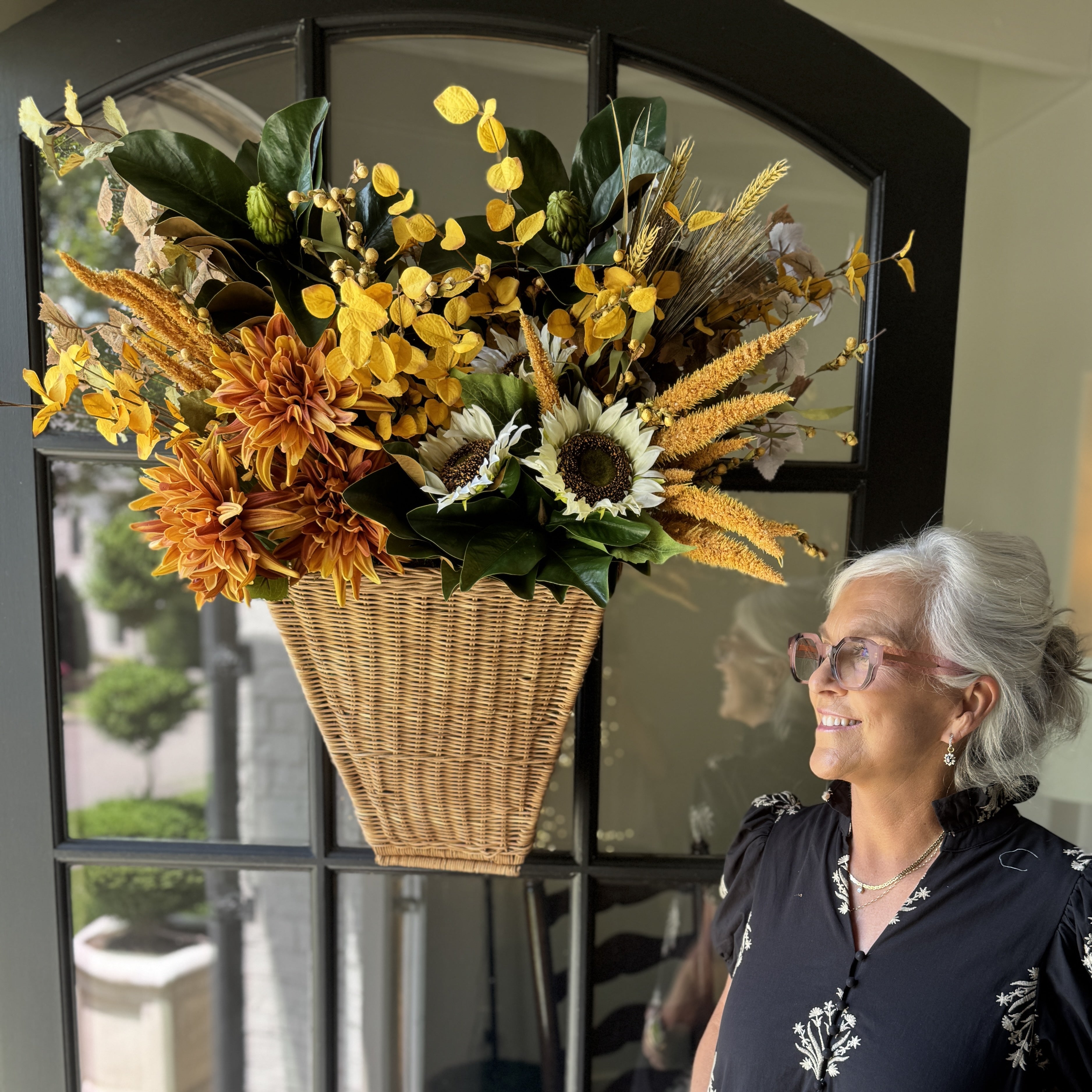 An older woman with gray hair and glasses stands indoors beside a dark gray door, smiling at a large autumn floral arrangement hanging on the door. The arrangement features yellow and orange flowers, sunflowers, wheat stalks, and green leaves in a woven basket.