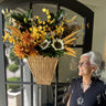 An older woman with gray hair and glasses stands indoors beside a dark gray door, smiling at a large autumn floral arrangement hanging on the door. The arrangement features yellow and orange flowers, sunflowers, wheat stalks, and green leaves in a woven basket.