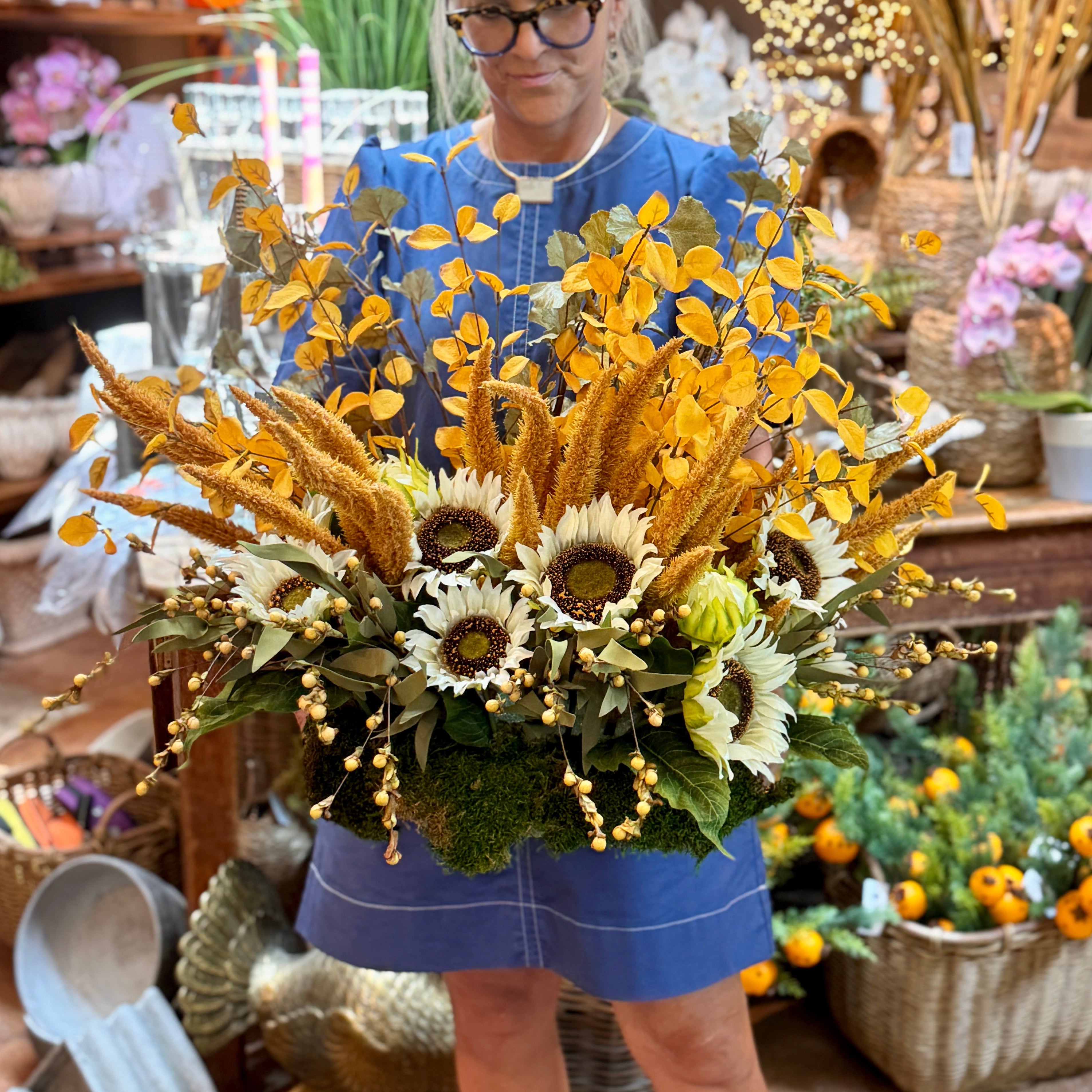 A woman with short gray hair and glasses stands in a rustic shop holding a large autumn floral arrangement featuring yellow sunflowers, wheat, and green foliage. She is wearing a blue dress, with shelves of fall decorations in the background.