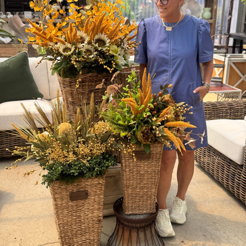 A woman in a blue dress stands beside three tall woven wicker flower baskets filled with autumnal floral arrangements in a garden center or patio setting.