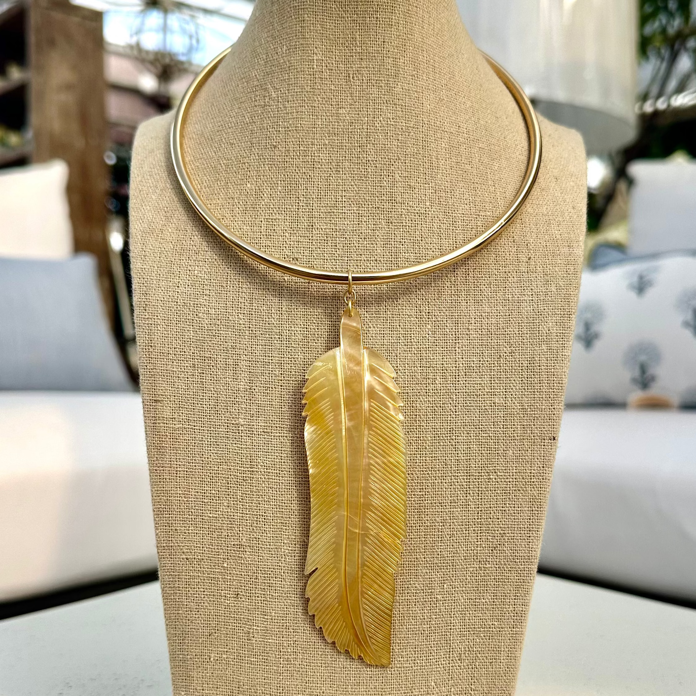 A hand-carved mother of pearl feather pendant on a gold-colored collar necklace, displayed on a mannequin bust.