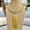 A hand-carved mother of pearl feather pendant on a gold-colored collar necklace, displayed on a mannequin bust.