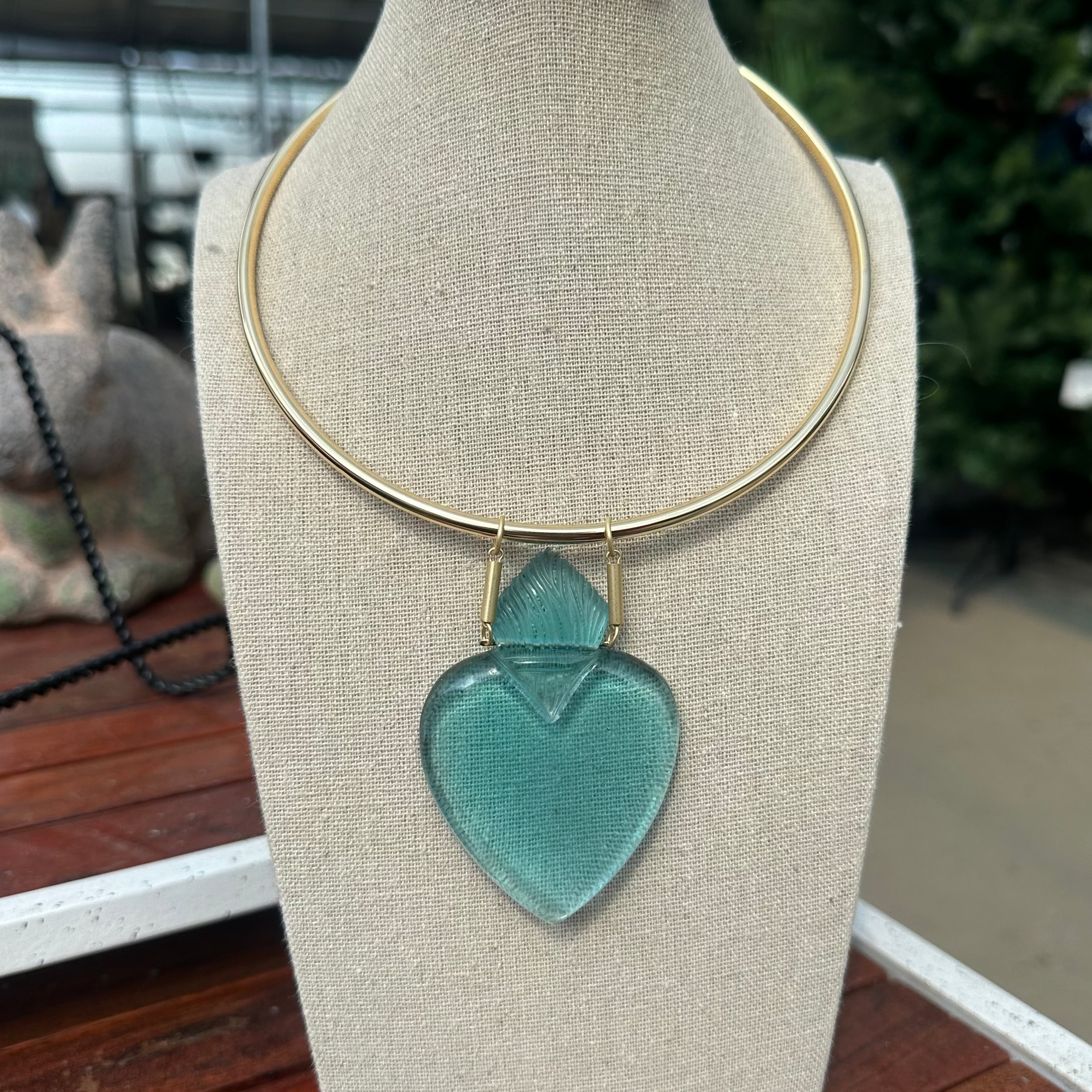 A gold-tone circular collar necklace with a translucent light blue-green heart-shaped glass pendant, displayed on a beige fabric mannequin bust.