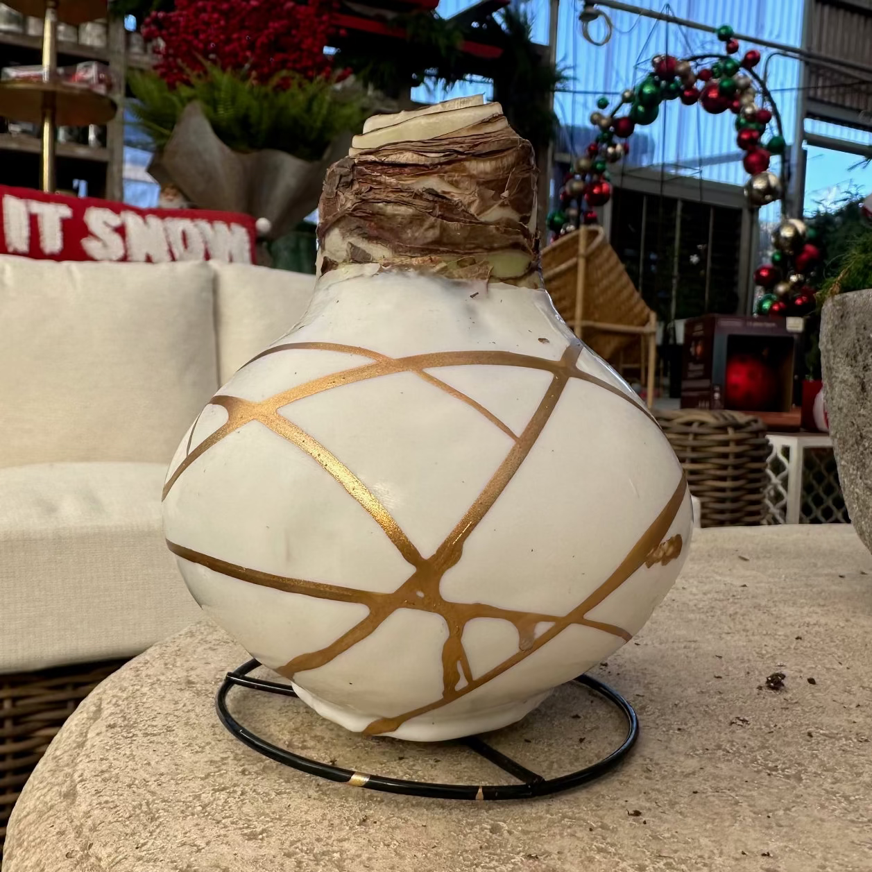 A white decorative Christmas ornament with gold painted lines and a brown textured neck, displayed on a stone table in a garden center or holiday decor store.