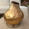 A metallic gold decorative vase with a crumpled foil texture and a dark brown top, displayed on a round stone table in an outdoor patio setting.