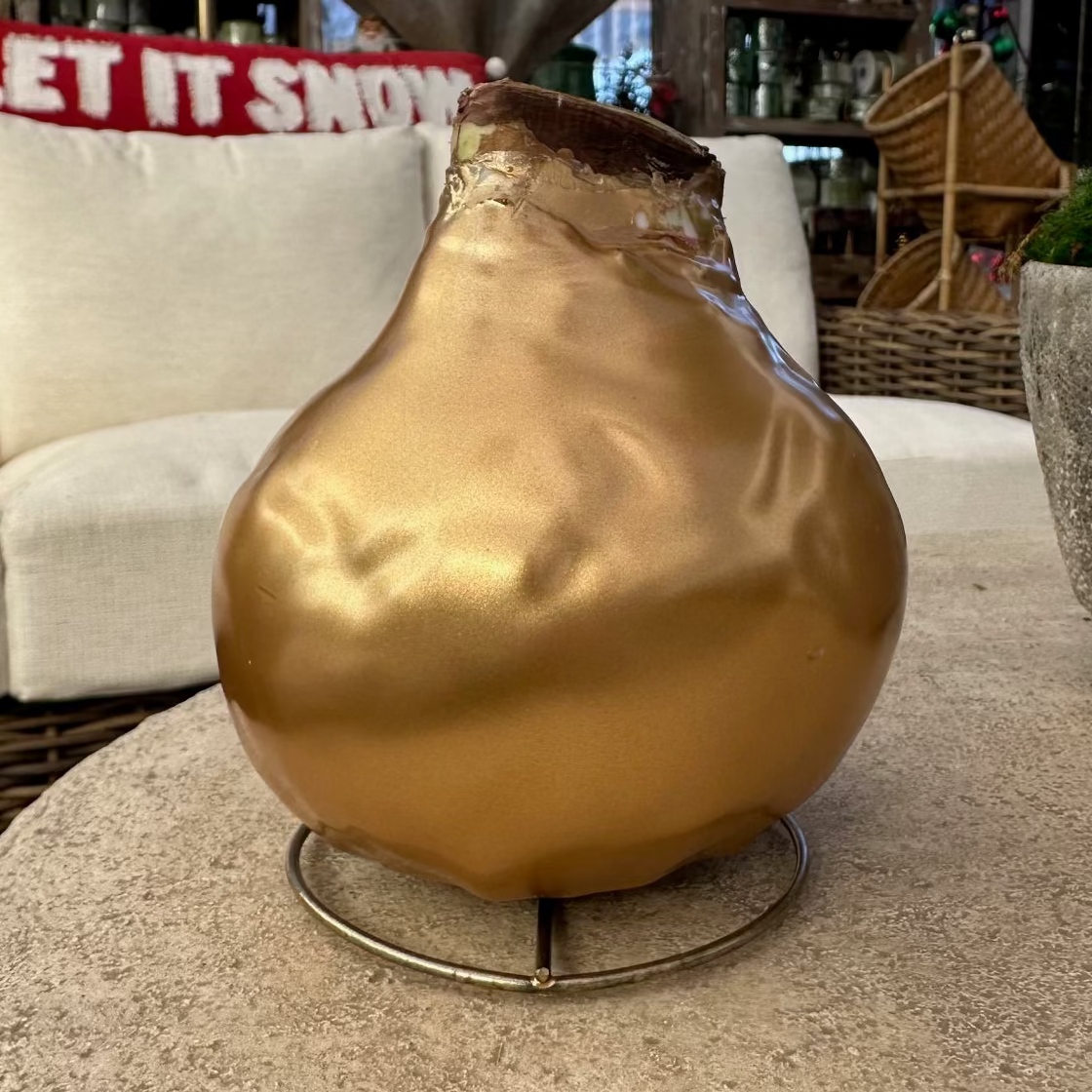 A metallic gold pear-shaped decorative vase with a small metal stand, displayed on a stone patio table in an outdoor patio setting with a white sofa and holiday decorations in the background.