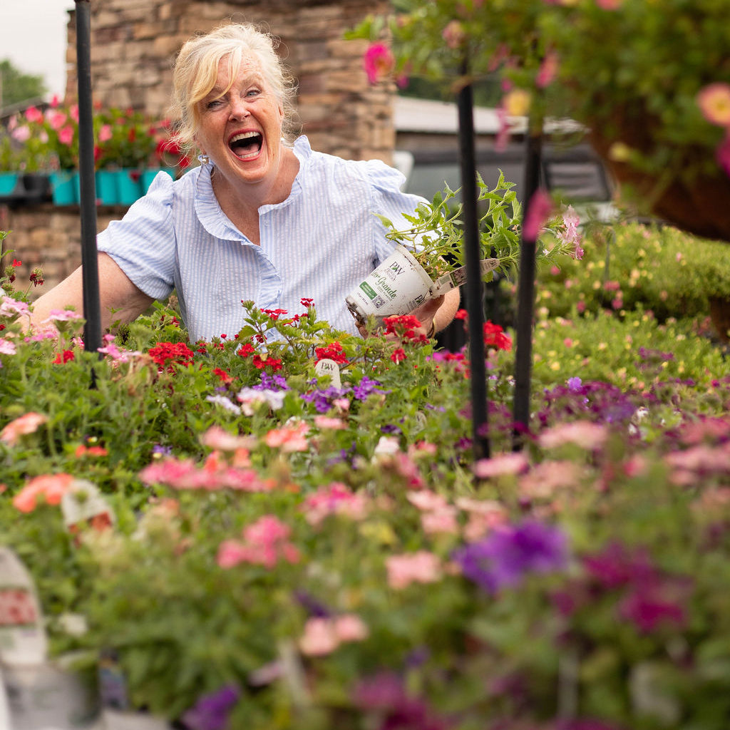 A woman with short blonde hair and glasses, wearing a light blue and white striped shirt, is smiling and laughing while holding a white plastic pot with a green plant in a garden center filled with colorful flowers.