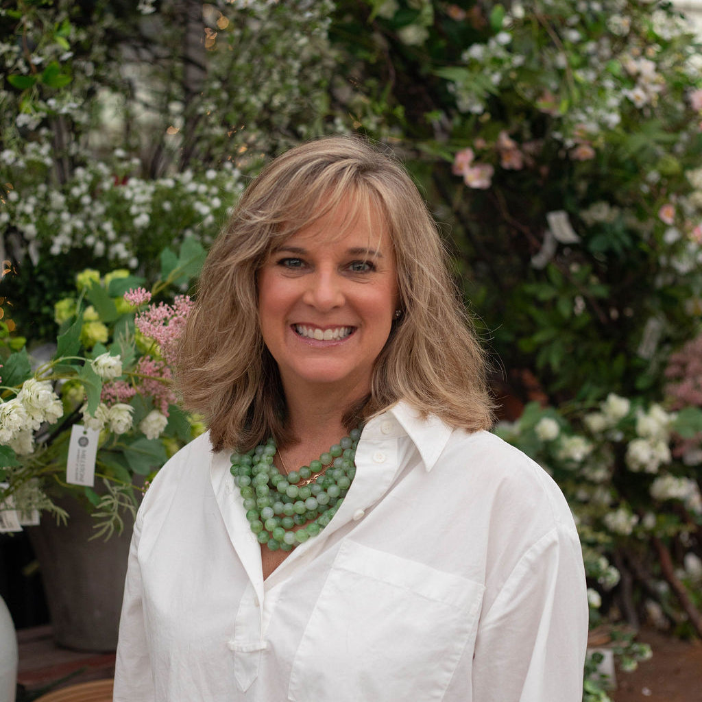 A smiling woman with blonde hair stands in a garden shop wearing a white button-down shirt and a chunky green beaded necklace, with lush green foliage and white flowers in the background.
