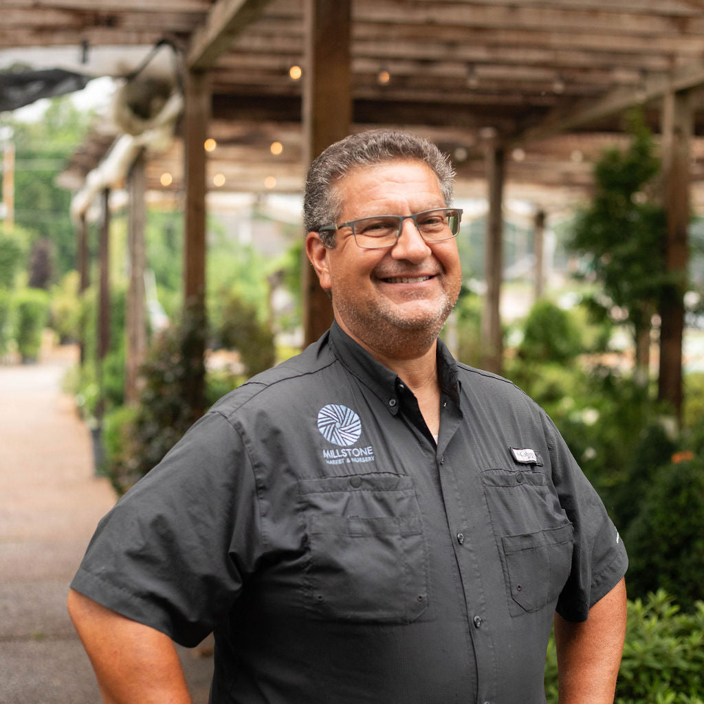 A smiling man wearing a dark gray short-sleeve button-up work shirt with a circular logo on the left chest stands under a wooden covered walkway with plants in the background.