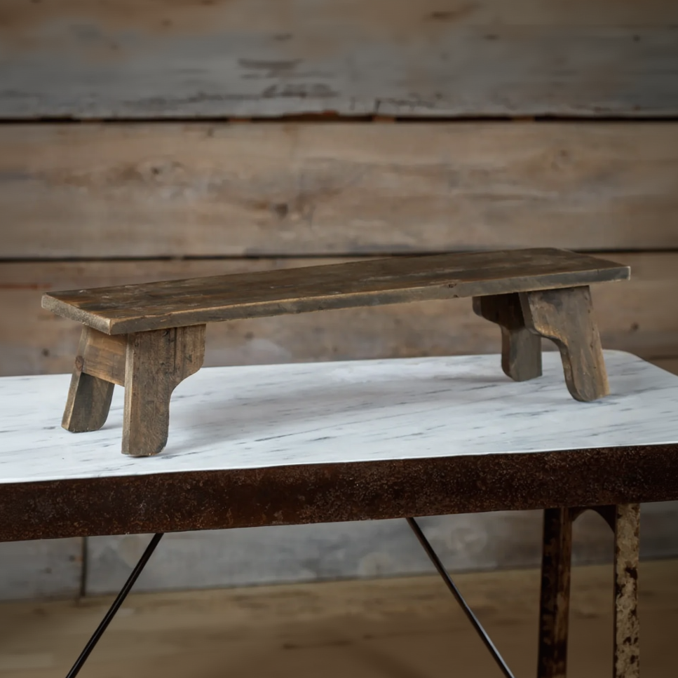 A rustic wooden bench with a weathered brown finish, shown resting on a white marble-topped table against a wooden plank wall background.