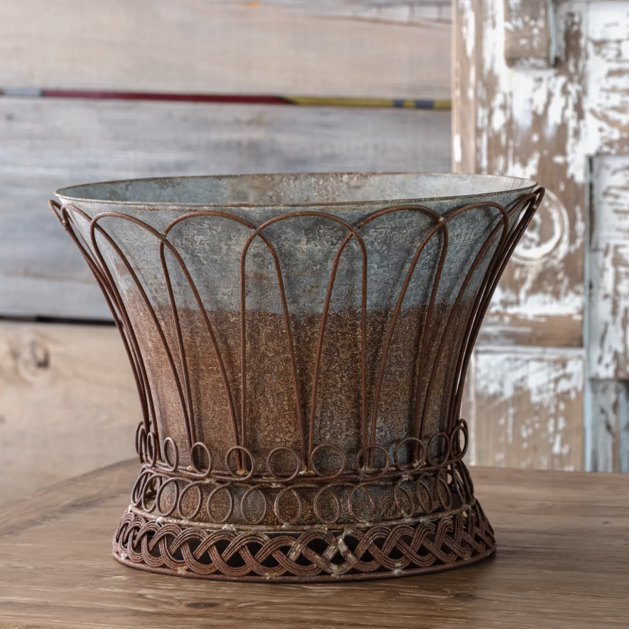 An ornate rusted metal planter with a scalloped rim and decorative circular wire base, displayed on a wooden surface against a rustic distressed wood background.