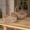 Two woven wicker baskets with tall handles, displayed on a wooden table in front of a house porch.