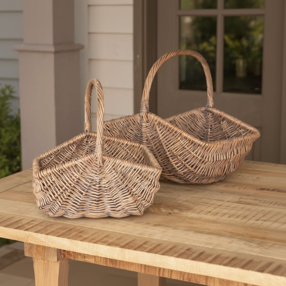 Two woven wicker baskets with tall handles, displayed on a wooden table in front of a house porch.