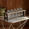 A rustic dark wood caddy holding six clear glass bottles, set on a weathered metal table against a wooden wall background.