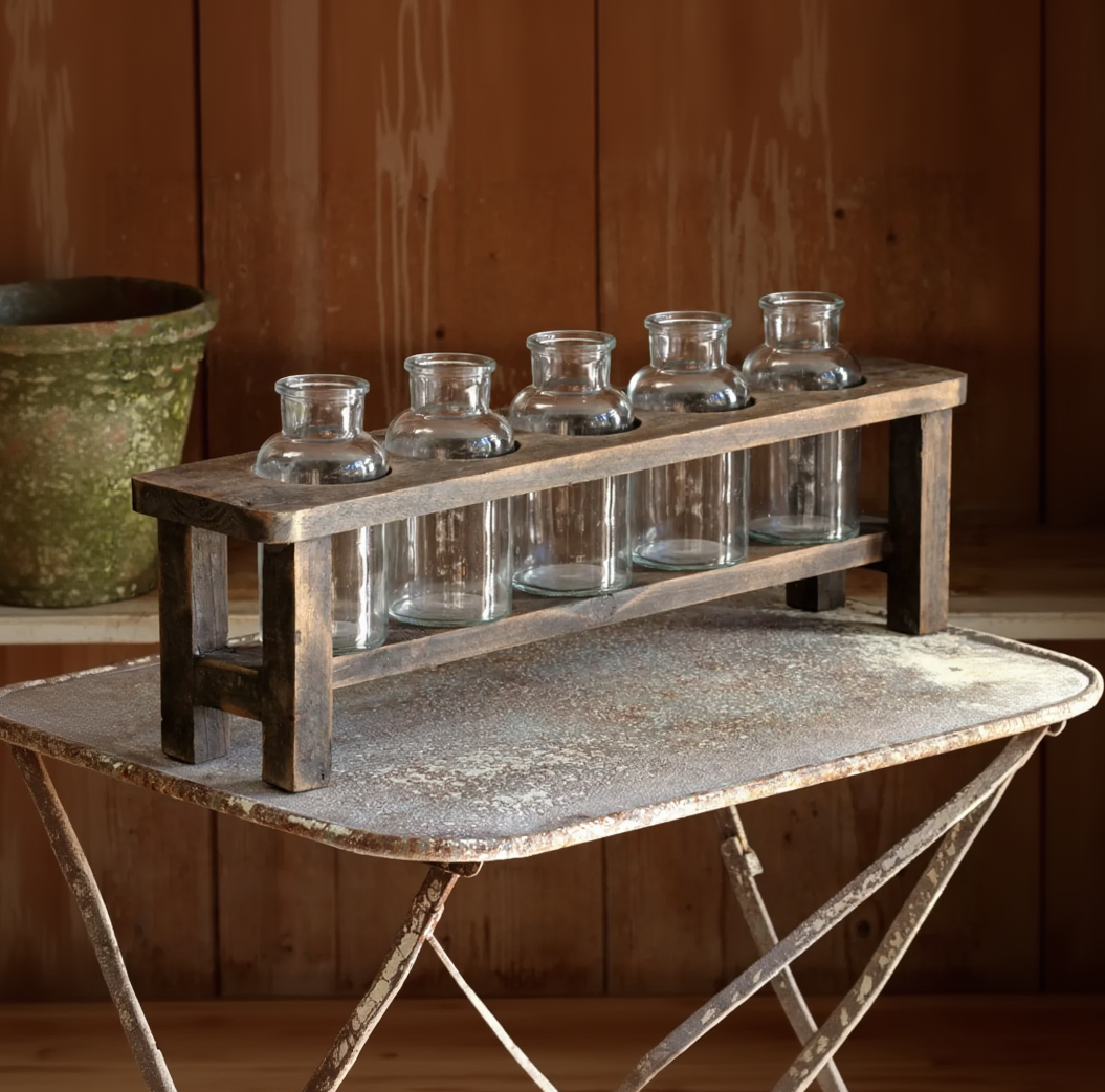 A rustic dark wood caddy holding six clear glass bottles, set on a weathered metal table against a wooden wall background.