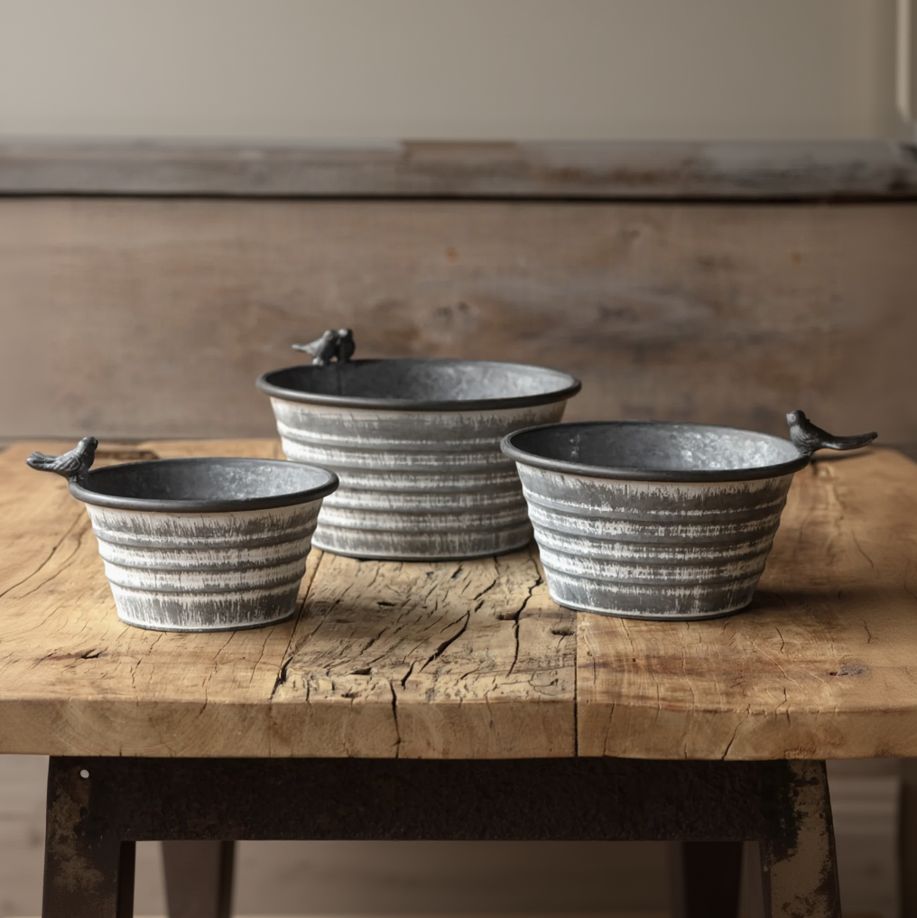 Three rustic galvanized metal bird feeder bowls of different sizes arranged on a wooden table against a wooden wall background.