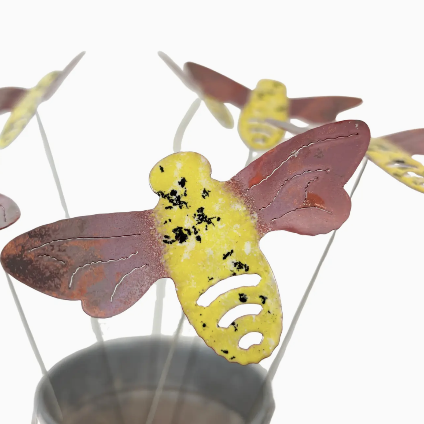 Metallic bee garden stakes with a central yellow and rust-colored bee ornament, surrounded by smaller matching bee decorations in a gray pot against a white background.