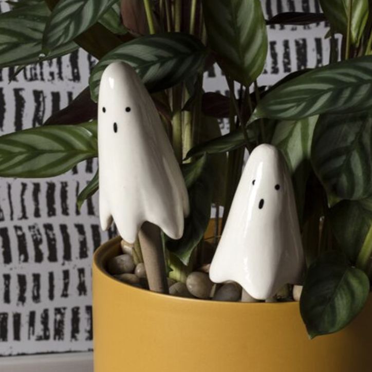 A yellow cylindrical planter holding a green leafy plant, with two small white ghost figurines placed among the leaves.
