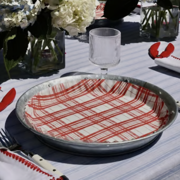 A round metal tray lined with a red and white plaid patterned fabric, set on a blue-and-white striped tablecloth with a glass, cutlery, and floral centerpieces.