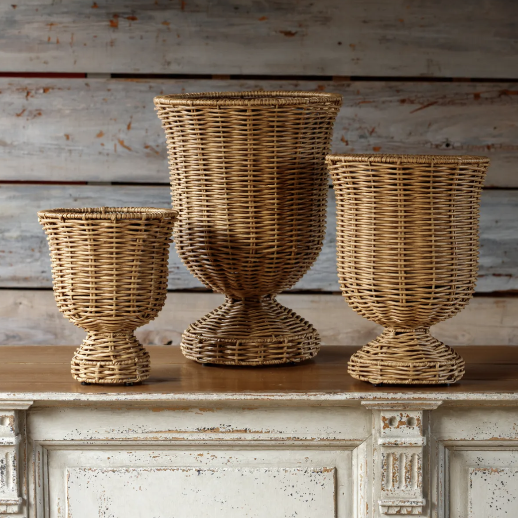 Three woven wicker planters in different heights, displayed on a distressed white wooden console table against a rustic gray wood plank wall.