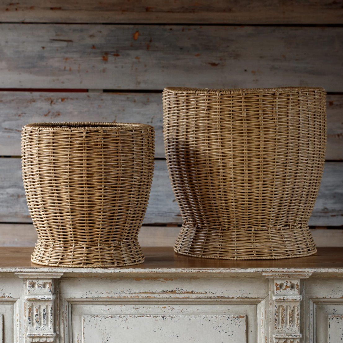 Two woven wicker planters in different sizes displayed on a distressed white wooden mantel against a rustic gray wood plank wall.