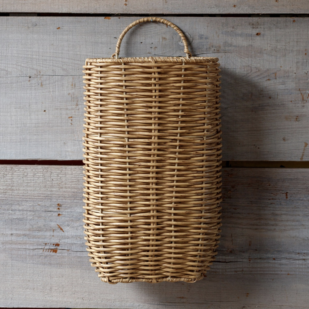 A natural woven wicker basket with a single top handle, displayed against a rustic, light gray wooden plank background.