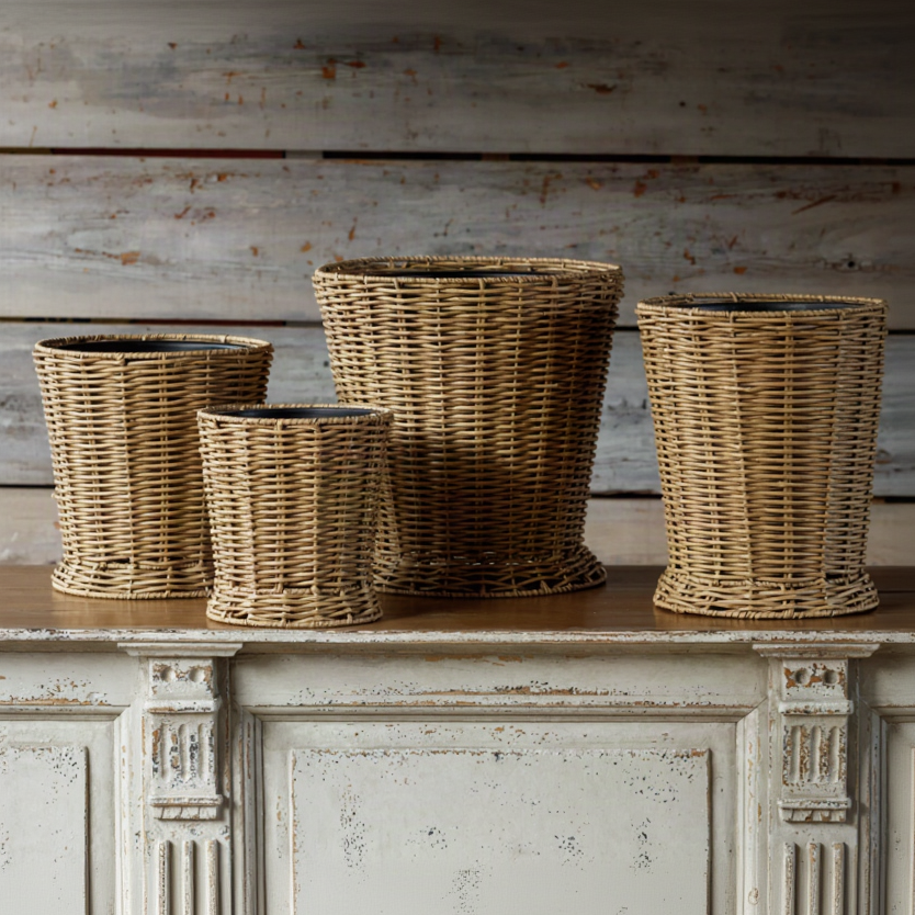 Four woven wicker planters in graduated sizes arranged on a distressed white wooden cabinet against a rustic weathered wood plank wall.