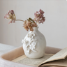 A white ceramic vase with embossed floral details sits on an open book inside a tray, holding dried pink flowers against a soft neutral background.