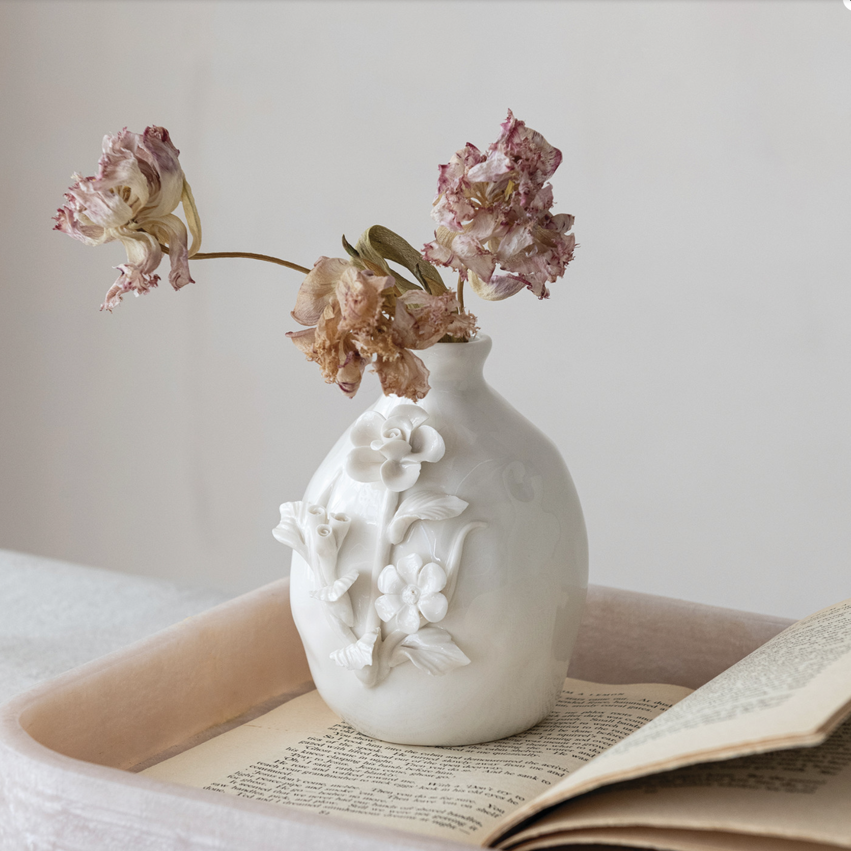 A white ceramic vase with embossed floral details sits on an open book inside a tray, holding dried pink flowers against a soft neutral background.