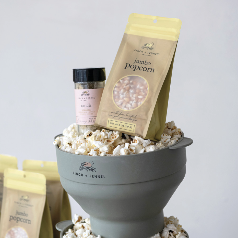 A gray funnel-shaped popcorn bowl filled with popcorn kernels and bags of Finch + Fennel jumbo popcorn, with a small jar of ranch seasoning in the background, photographed on a light gray studio background.
