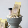 A gray funnel-shaped popcorn bowl filled with popcorn kernels and bags of Finch + Fennel jumbo popcorn, with a small jar of ranch seasoning in the background, photographed on a light gray studio background.