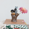 A small bird figurine perched on a tree stump, holding a pink flower, displayed on top of a tan book with a patterned textile underneath against a light gray background.