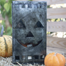 A rustic metal jack-o'-lantern lantern with a distressed dark gray finish, styled on a bed of straw with small pumpkins in a garden setting.