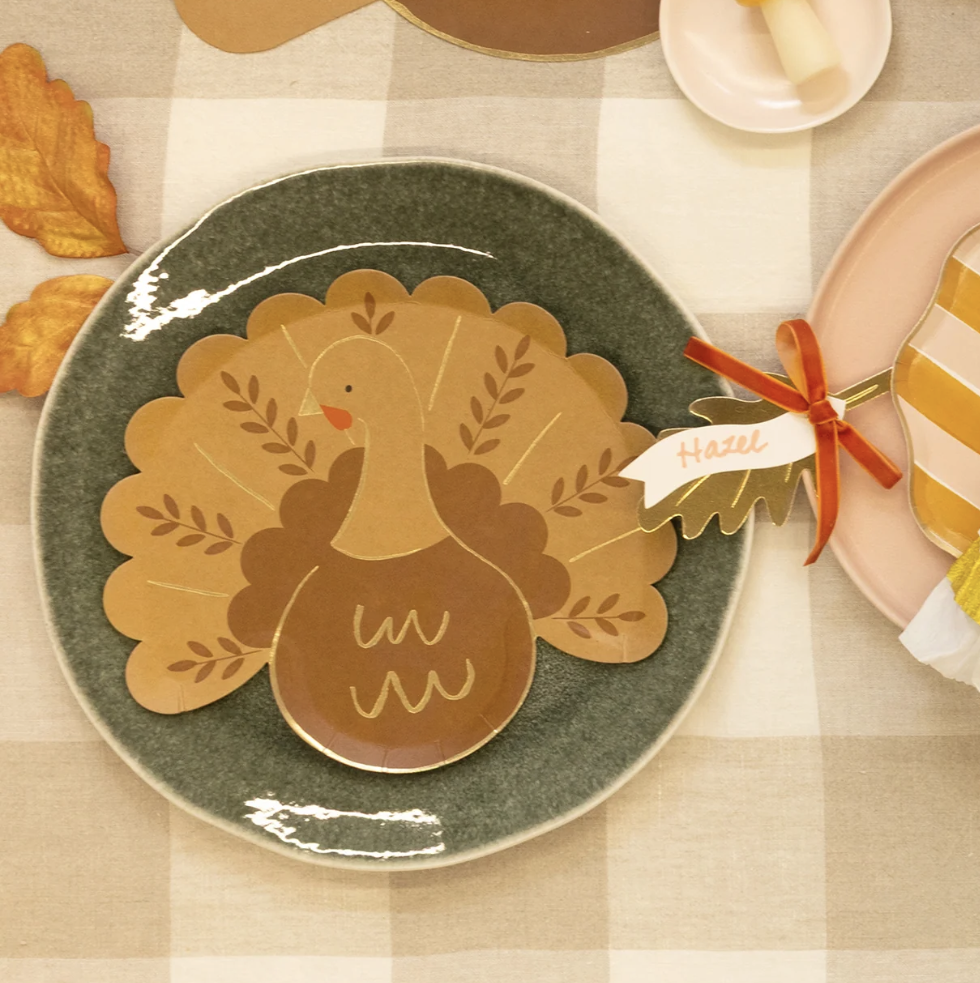 A styled autumn table setting featuring a ceramic plate with a turkey-shaped placemat, a striped napkin tied with a red ribbon, pumpkins, fall leaves, and a checkered tablecloth.