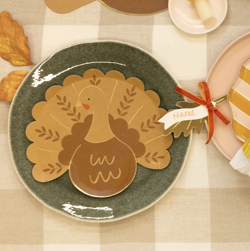 A styled autumn table setting featuring a ceramic plate with a turkey-shaped placemat, a striped napkin tied with a red ribbon, pumpkins, fall leaves, and a checkered tablecloth.