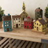 A set of hand-painted wooden house ornaments shaped like a small village, including a church, colorful houses, and a car, displayed on a wooden shelf.