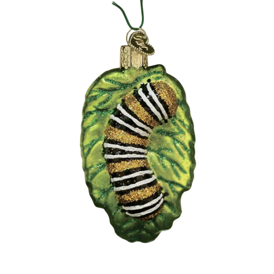 A green leaf-shaped Christmas ornament hanging on a tree, featuring a glittery caterpillar design in black, white, and gold stripes against a softly blurred holiday background with warm lights.