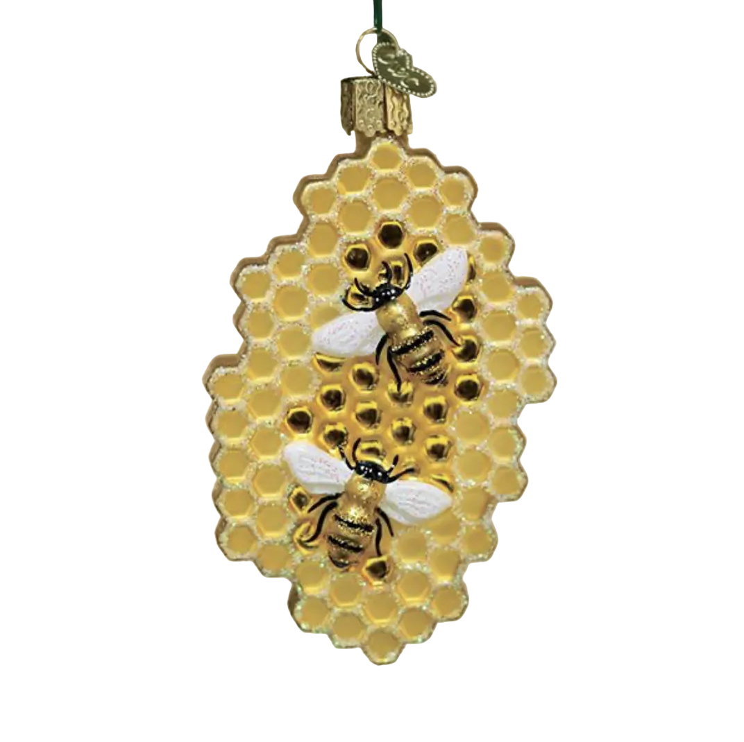 A decorative hanging ornament shaped like a honeycomb with two detailed bees on it, shown hanging in front of a softly blurred Christmas tree and warm bokeh lights.