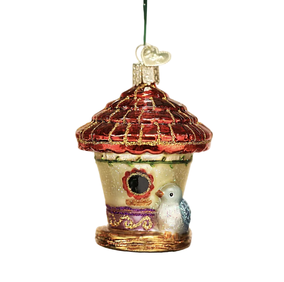 A decorative Christmas birdhouse ornament hanging from a green pine branch. The ornament features a red glittery roof, a cream-colored body, purple and gold trim, and a small white bird with blue accents standing on the front.