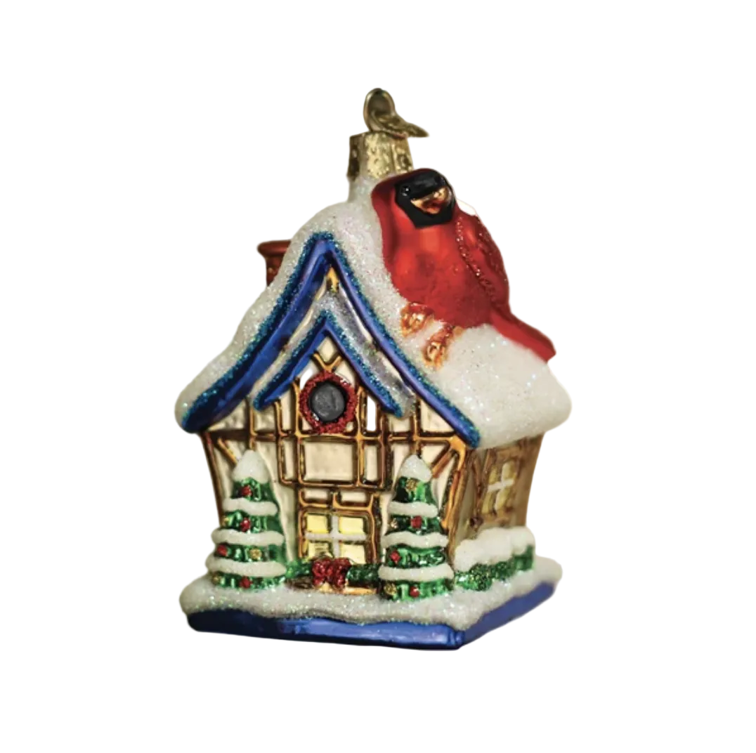 A Christmas house ornament hanging on a tree branch, featuring a red bird on a snow-covered roof, with warm blurred lights in the background.