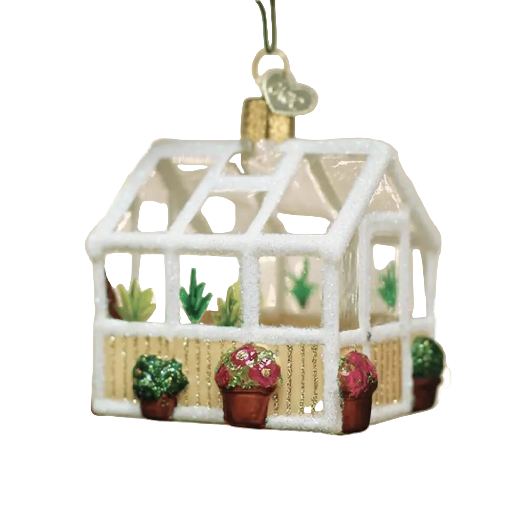 A white-framed greenhouse Christmas ornament with small potted plants, hanging from a green tree branch against a warm, blurred holiday background.