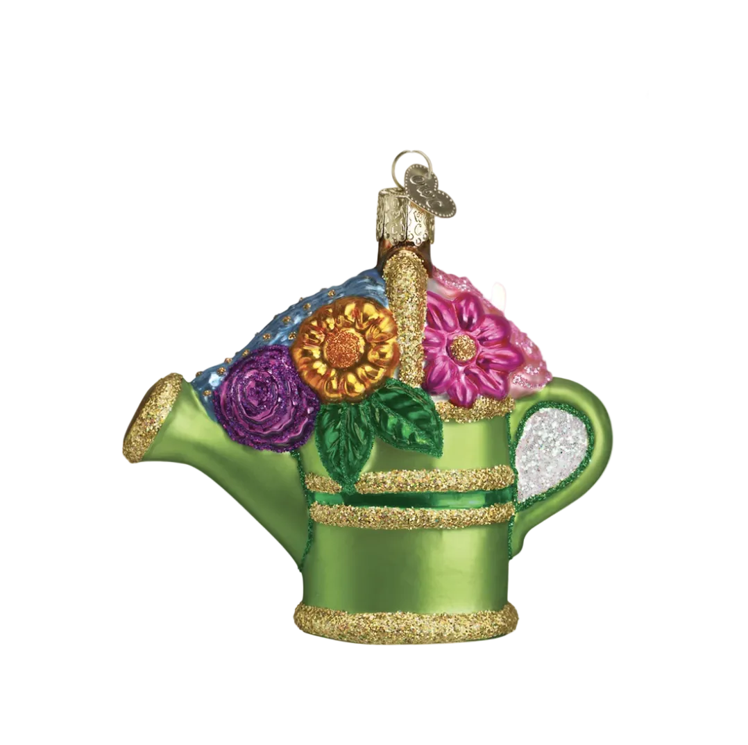 A green watering can Christmas ornament decorated with colorful faux flowers and gold glitter accents, hanging from a pine tree branch against a warm, softly blurred background with bokeh lights.