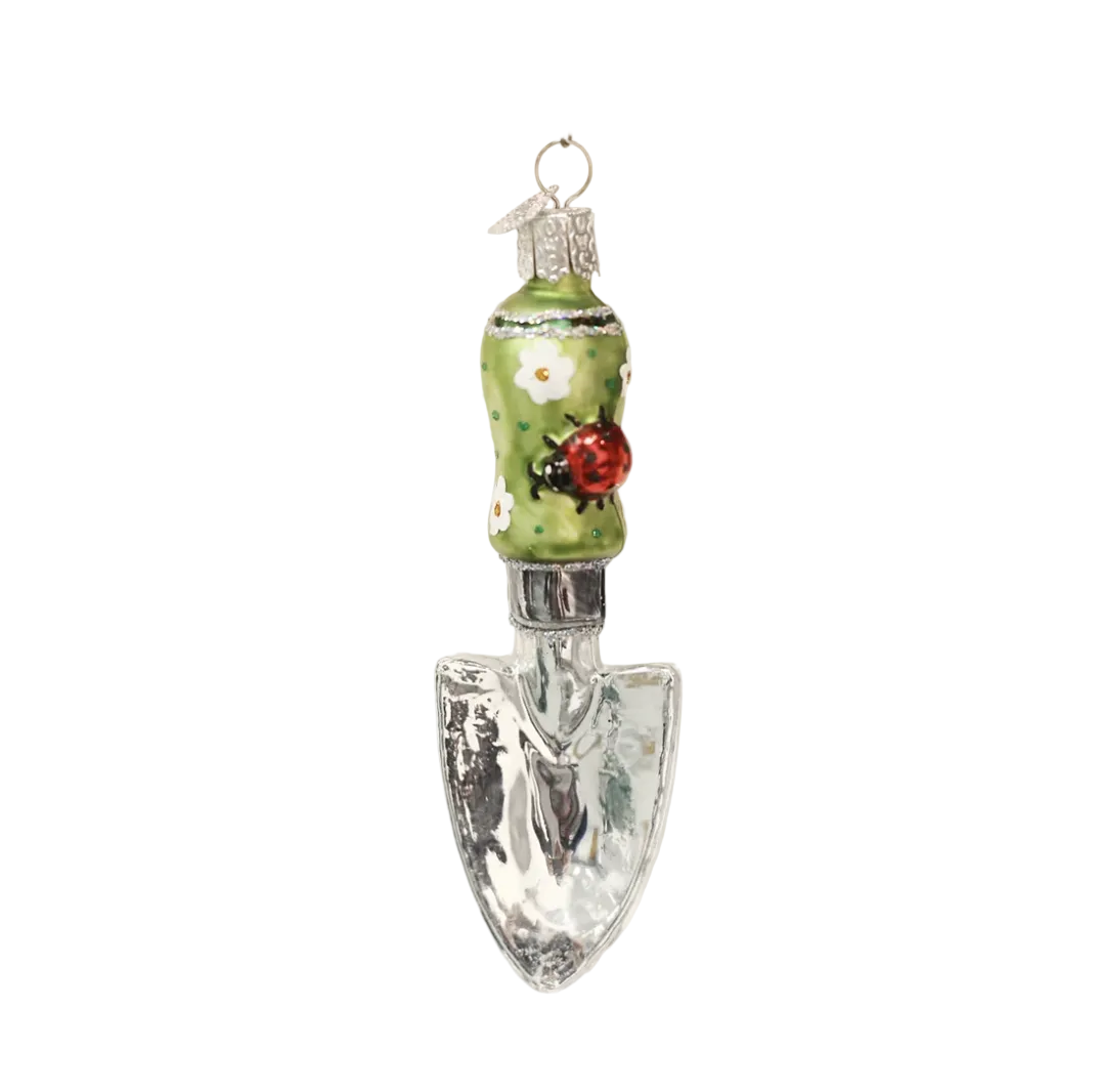 A Christmas ornament shaped like a small hand trowel hanging from a pine tree branch. The trowel handle is green with white daisies, a red ladybug, and silver glitter, while the blade is clear glass.