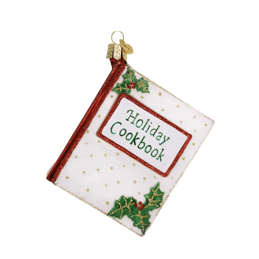 A Christmas ornament shaped like a cookbook, hanging from a tree branch. The cookbook ornament is white with gold glitter dots, a red glitter spine, and green holly accents, with the words 'Holiday Cookbook' written on the front.