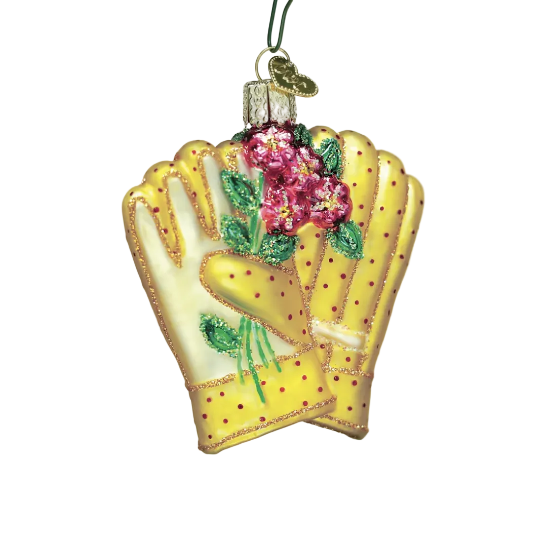 A decorative Christmas ornament shaped like a yellow baseball glove with pink flowers and green leaves, hanging from a tree branch with blurred holiday lights in the background.