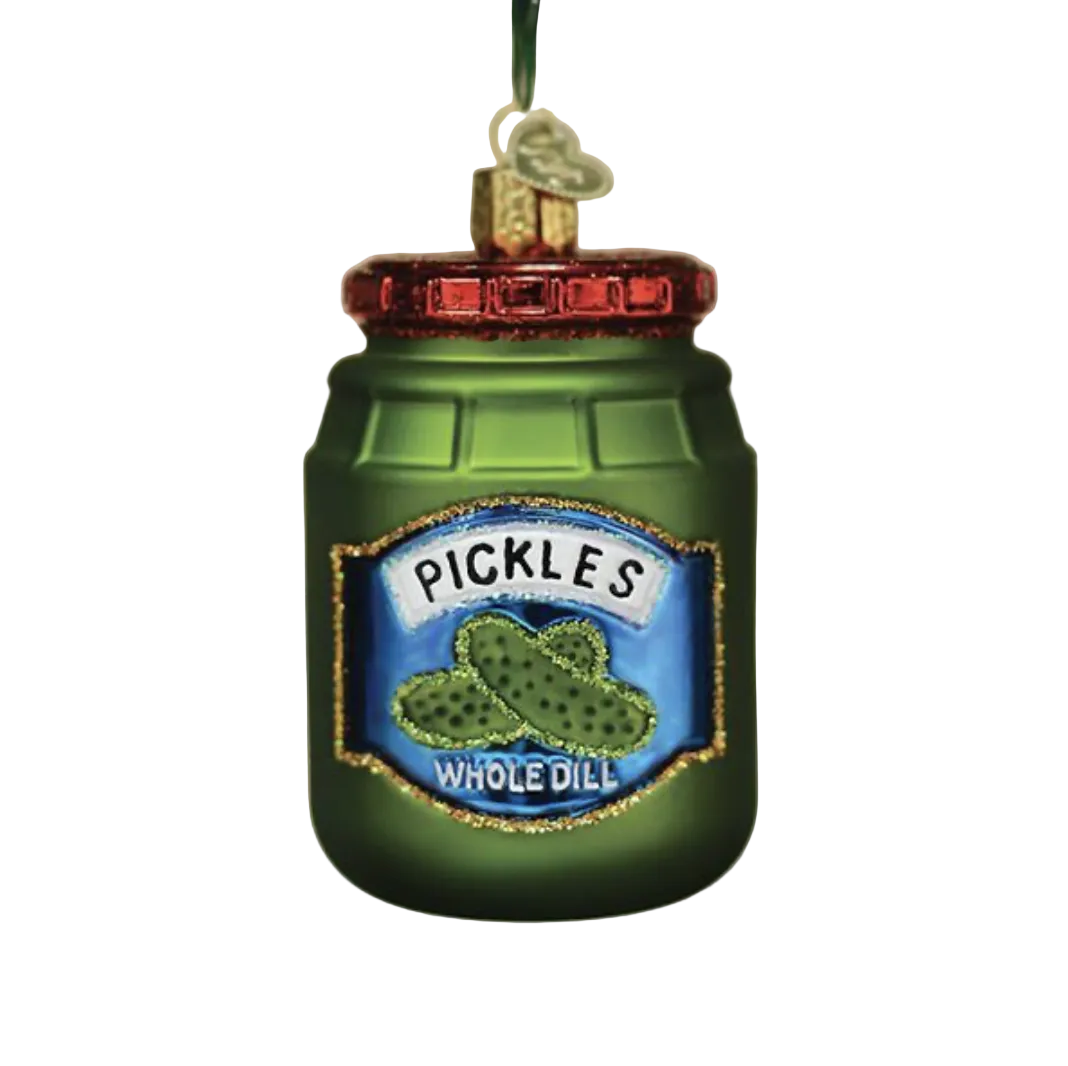 A green pickle jar Christmas ornament hanging from a tree branch, with a blue 'Pickles Whole Dill' label and a red lid, against a softly blurred holiday background.
