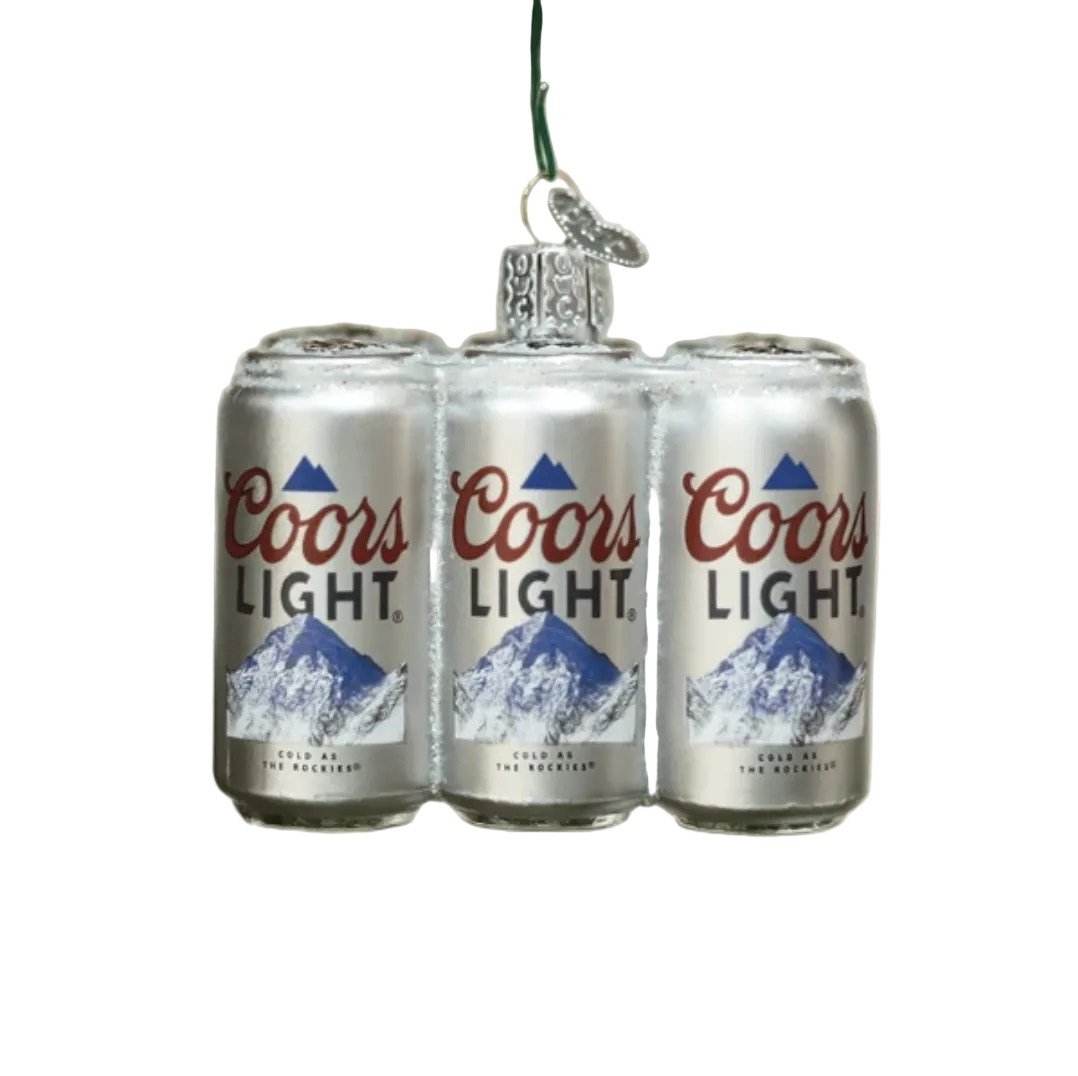 A Coors Light Christmas ornament shaped like three silver beer cans hanging from a green string, surrounded by pine branches and warm white bokeh lights in the background.