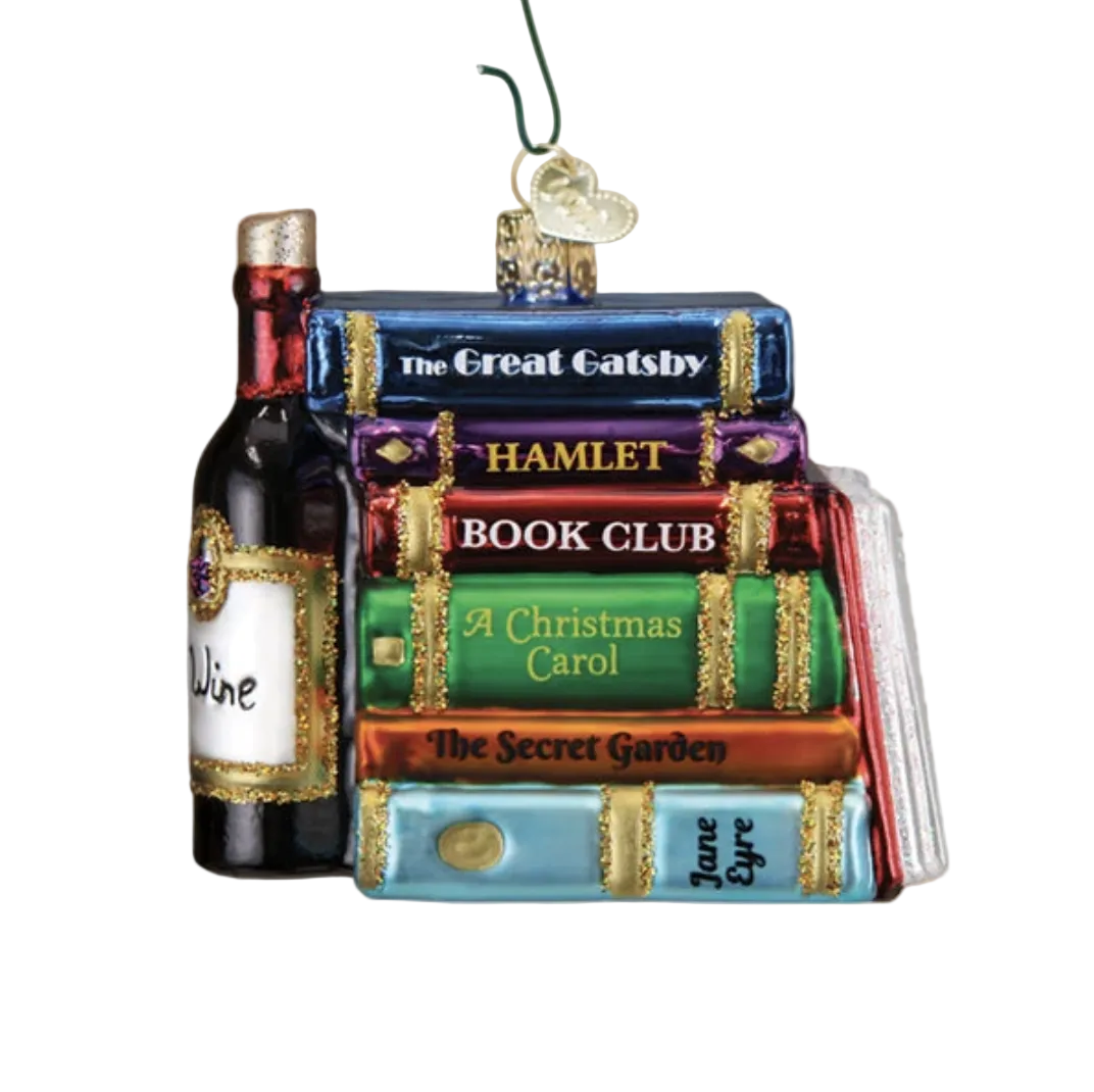 A Christmas ornament shaped like a stack of books with a wine bottle on the left, hanging from a green hook. The ornament features titles such as The Great Gatsby, Hamlet, Book Club, A Christmas Carol, The Secret Garden, and Jane Eyre, with a warm blurred holiday background and tree branches in the foreground.